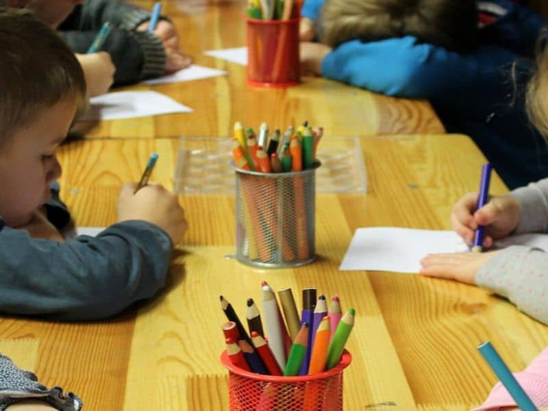 Children sitting at a wooden table, drawing with colored pencils in small metal containers. Focus on creativity and concentration in a classroom setting.