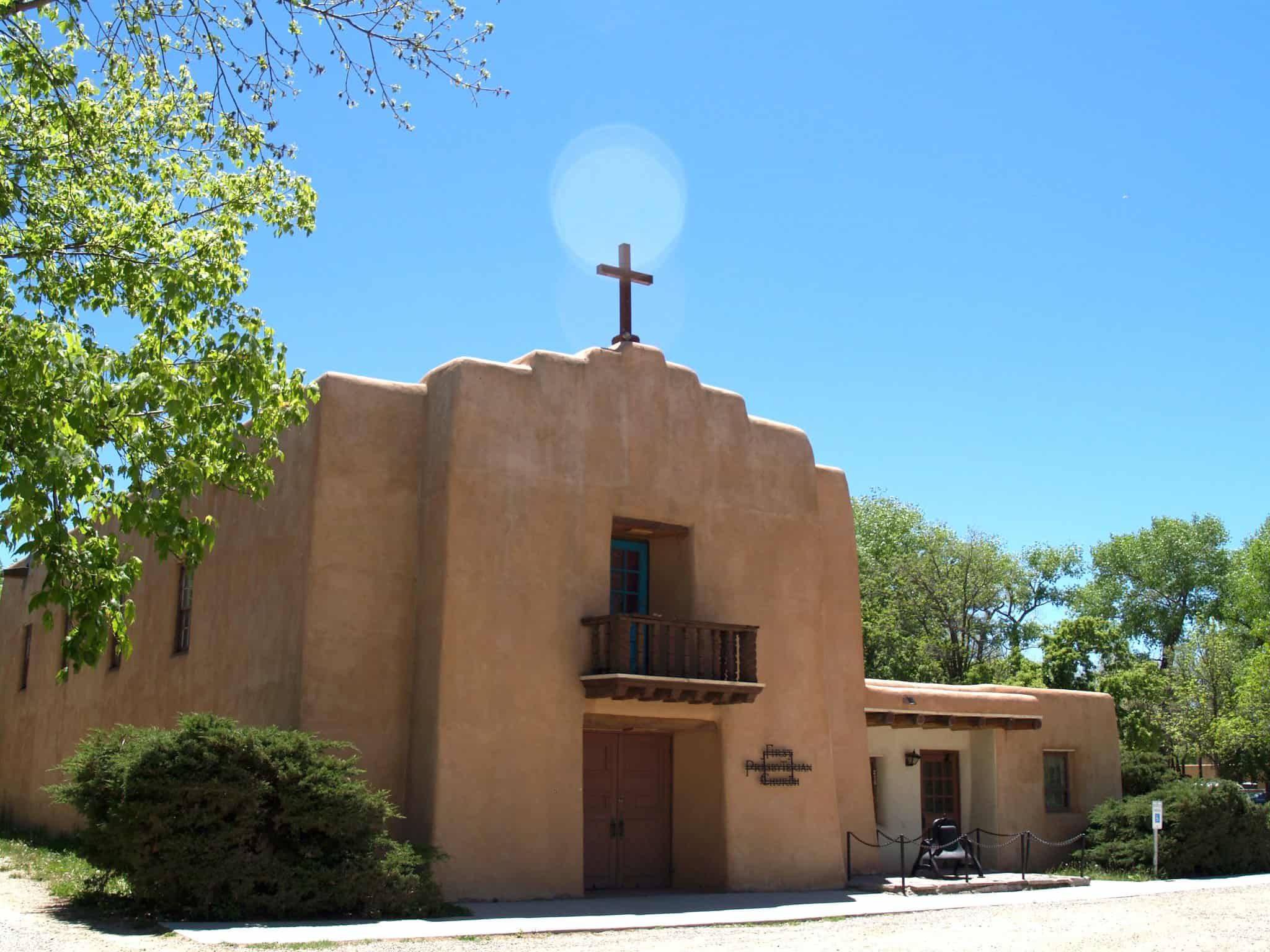 Adobe-style building with cross and balcony, surrounded by greenery under a clear blue sky. Front sign reads "First Presbyterian Church." Person standing nearby.