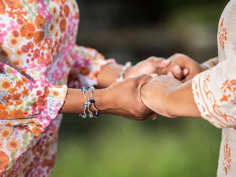 Two people in colorful patterned clothing hold hands, displaying bracelets against a blurred green background, symbolizing connection and support.