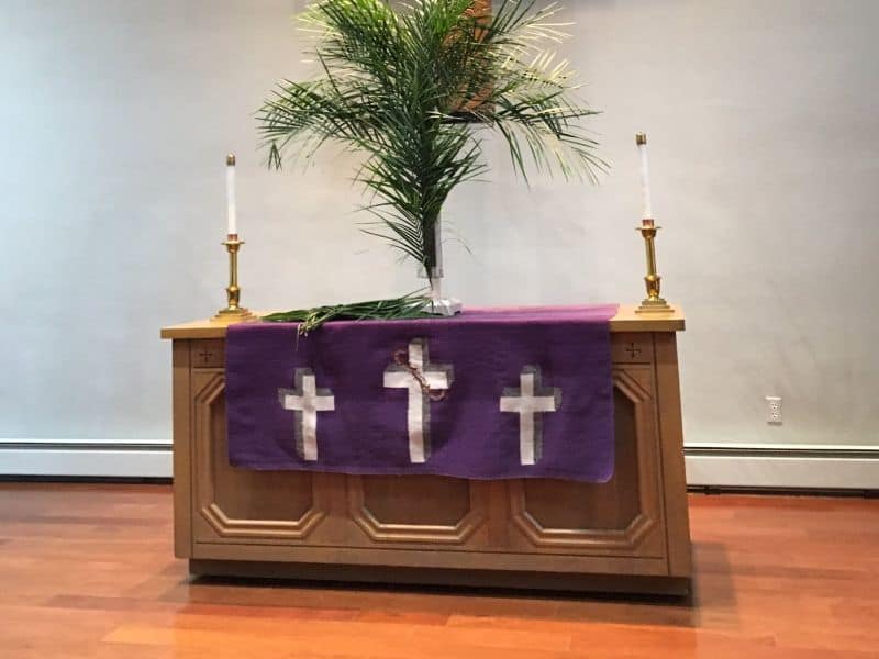 An altar with palm branches, two candles, and a purple cloth adorned with crosses, set against a wooden floor and plain background.