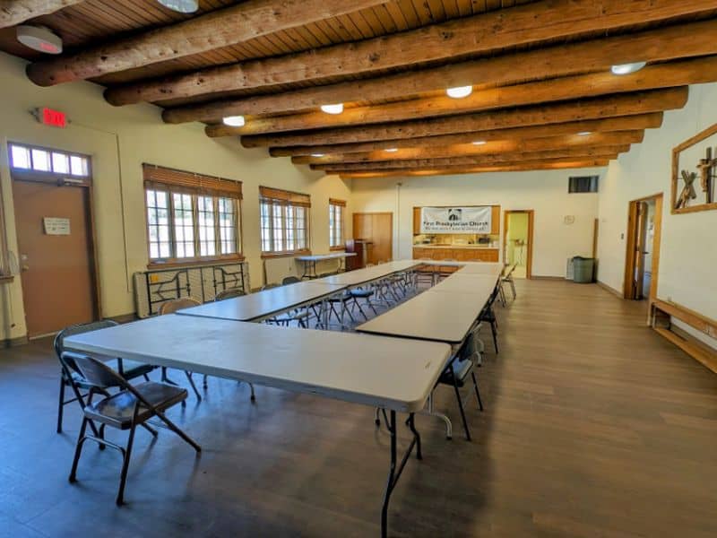 A spacious meeting room with wooden beams, long tables, folding chairs, and a banner reading "First Presbyterian Church" on the wall.