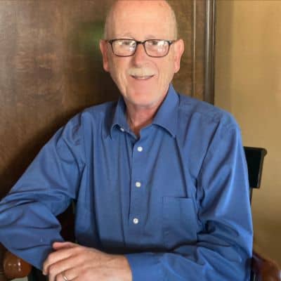 A person wearing glasses and a blue shirt is sitting indoors, smiling at the camera. The background features a wooden door.
