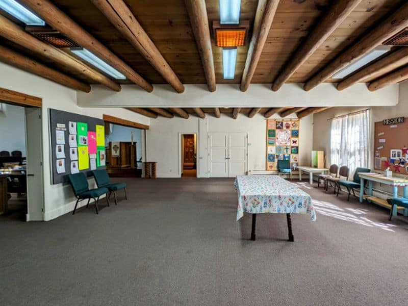 Spacious room with wooden beam ceiling, colorful posters on walls, and a table in the center. Chairs are arranged along the walls.