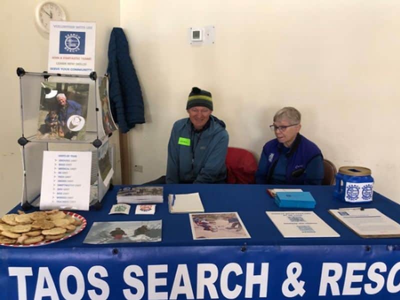 Two people sit at a Taos Search & Rescue booth with brochures and cookies on a table.
