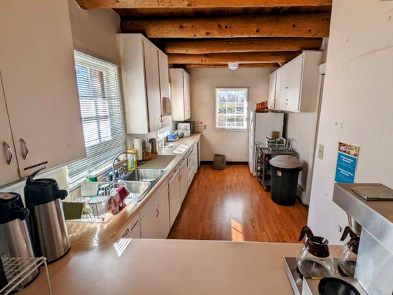 A cozy kitchen with wooden beam ceiling, white cabinets, stainless steel sink, and coffee station. Natural light from two windows brightens space.