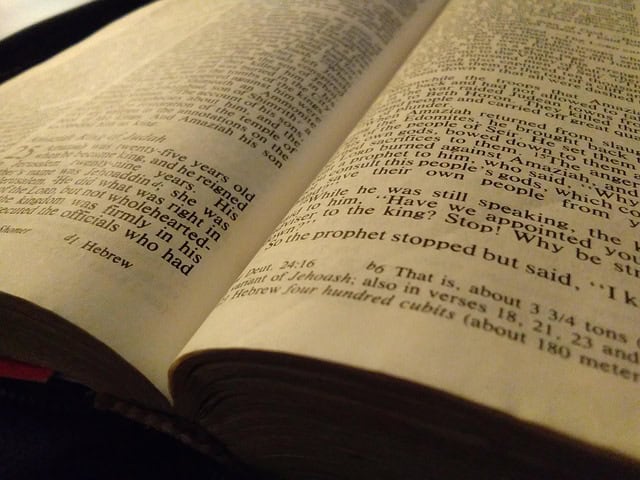 An open Bible displaying paragraphs and verses in English, resting on a dark surface with a visible crease along the spine.