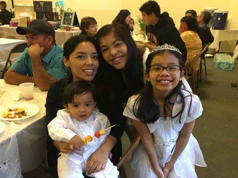 A smiling group at an indoor gathering, featuring a person holding a child with food on a skewer. Others are seated nearby.