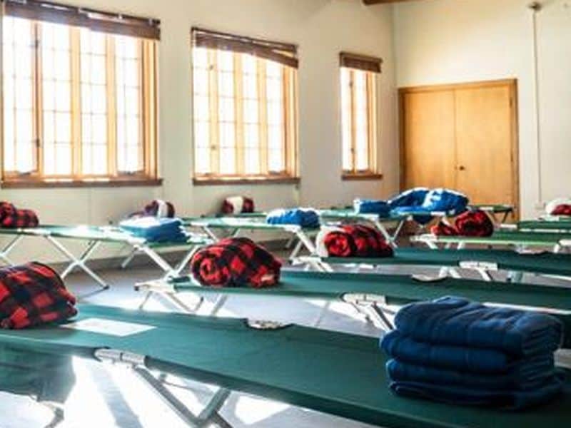 The image shows a room with rows of cots, each with folded blankets. Sunlight streams through large windows, brightening the space.