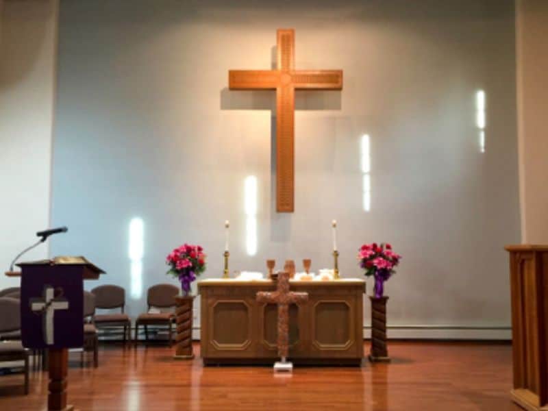 A church altar with a large cross, vibrant flowers, candles, lectern, and chairs. Warm lighting enhances the peaceful ambiance.