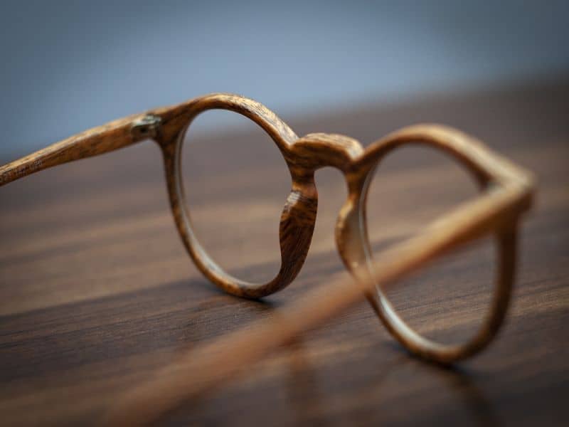 Wooden eyeglasses with a round frame sit on a wooden surface, showcasing a rustic aesthetic against a soft, blurred background.