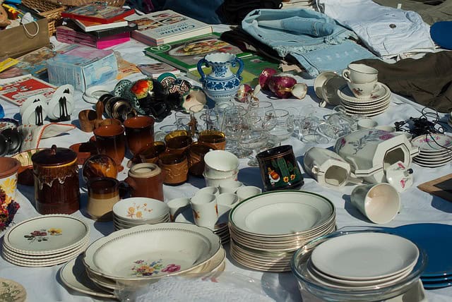 A variety of vintage dishes, glasses, and ceramics are displayed on a table at an outdoor market, surrounded by books and clothing.