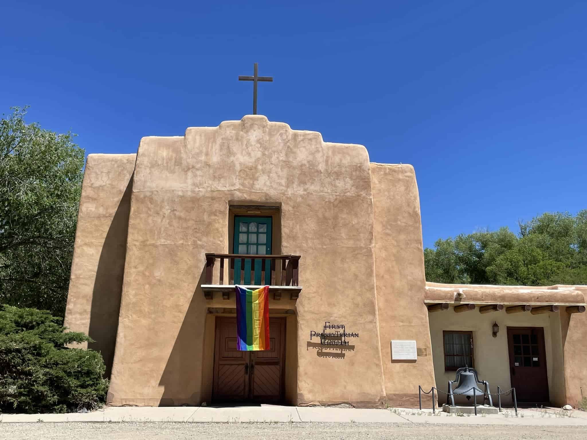 A brown adobe church with a cross on top, rainbow flag, and a large bell outside. Clear blue sky and trees in background.
