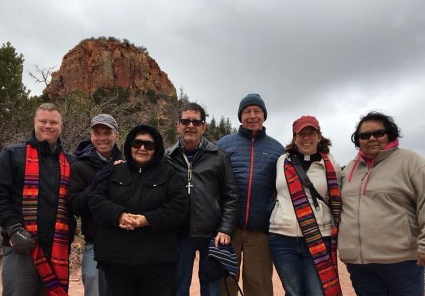 A group of seven people pose outdoors, dressed warmly, with a rocky hill and trees in the background under a cloudy sky.