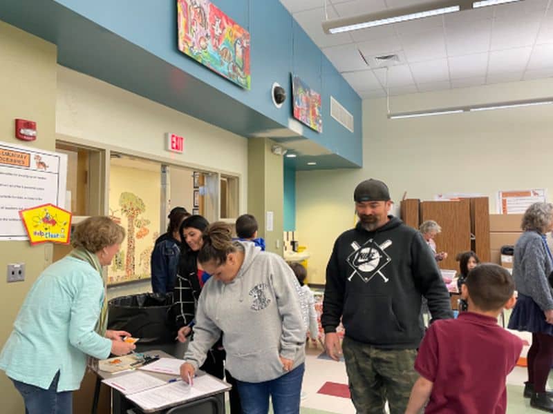 People and children engage in activities at a community center. Colorful artwork decorates the walls. There are tables, chairs, and informational signs.