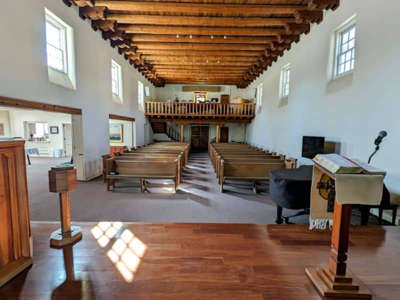 The image shows an empty church interior with wooden pews, pulpit, and balcony. Light streams through windows, highlighting the wooden ceiling beams.