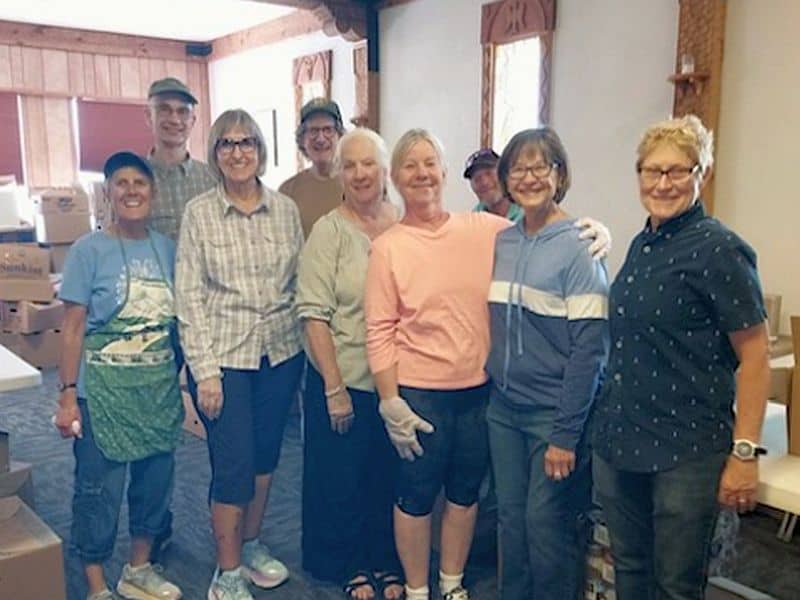 A group of nine smiling people posing indoors, with cardboard boxes in the background, suggesting a community or volunteer activity setting.