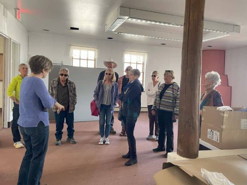 A group of people is attentively gathered in a plain room with wooden beams and boxes, engaged in conversation or a presentation.
