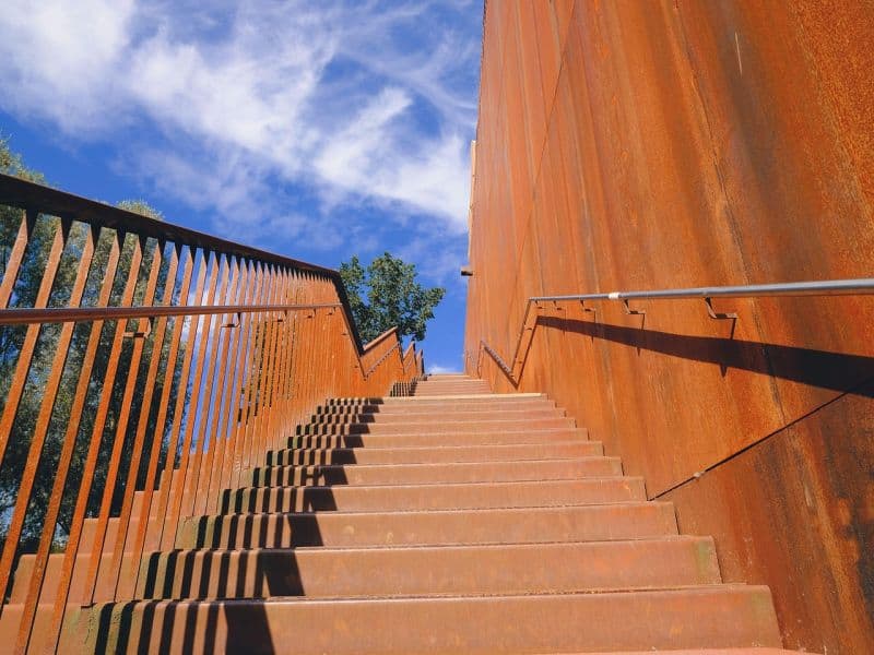 Rust-colored outdoor staircase beside a metal wall under vibrant blue sky, leading upwards with trees visible at the top.