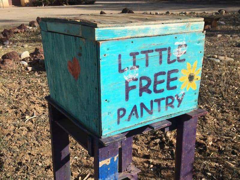 A turquoise wooden box labeled "Little Free Pantry" with a heart and flower design, placed outdoors on a purple stand.