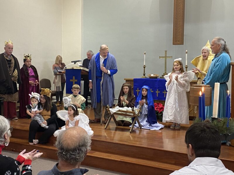 People and children in costumes perform a nativity scene in a church. Candles, crosses, and blue decorations are visible on the altar.