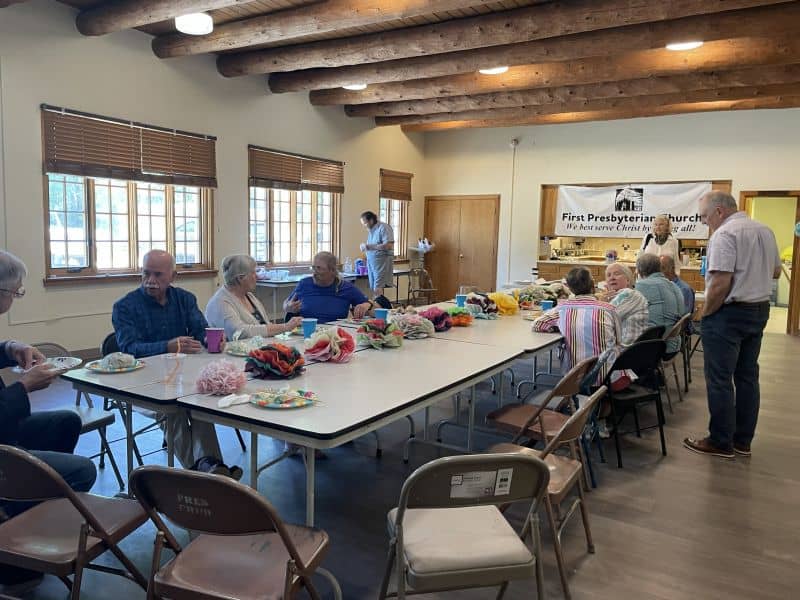 A group of people gathers in a room with a wooden ceiling at First Presbyterian Church. Colorful items are arranged on tables.