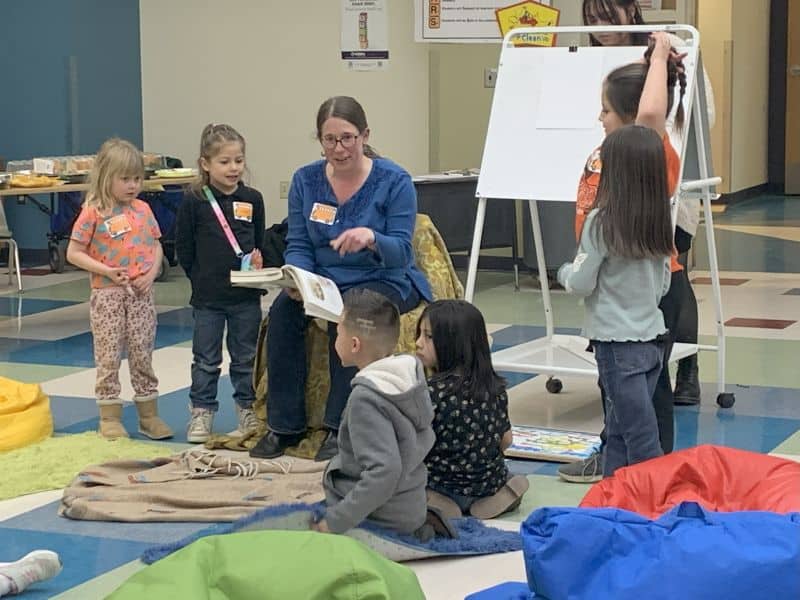 A person reads to several children seated around them in a colorful room with a whiteboard and bean bags.