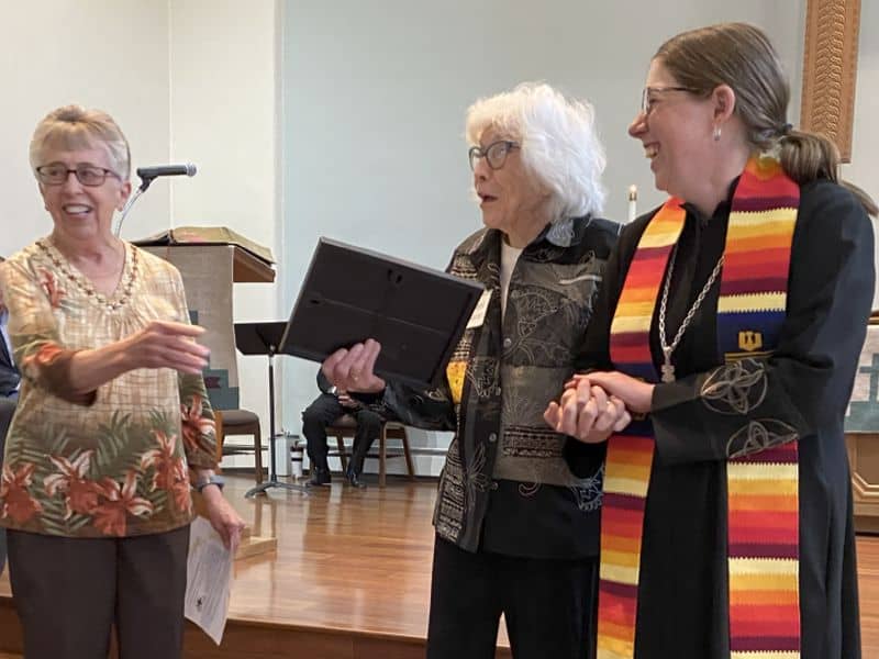 Three people gather indoors, conversing and smiling, with a microphone and chairs in the background. One person wears a multicolored stole.