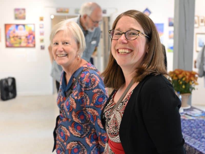 Two people smile inside an art gallery, with colorful paintings on the walls. A person in the background observes the artwork.