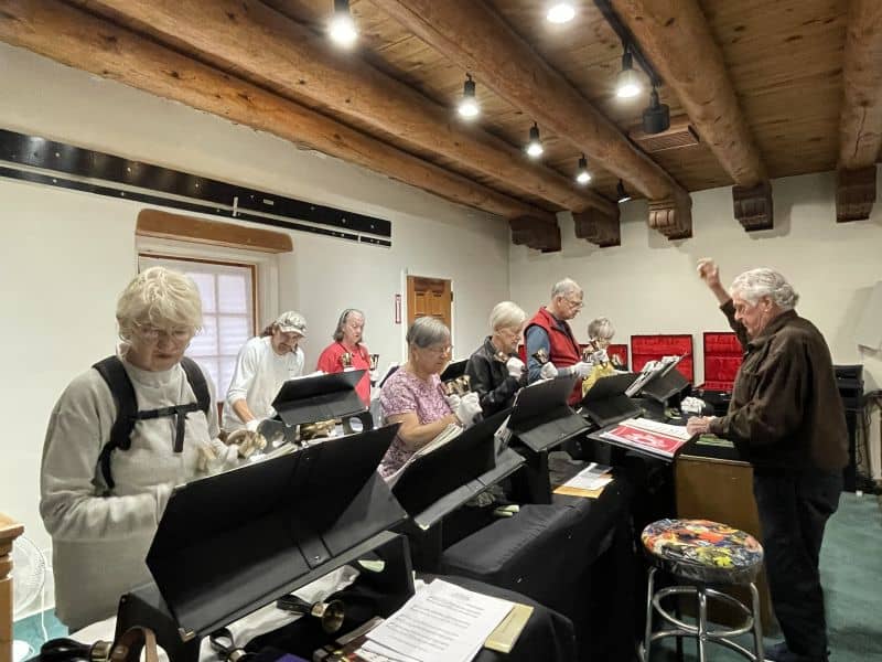 A group of people are playing handbells in a room with wooden beams. They are focused on their music sheets and conductor.