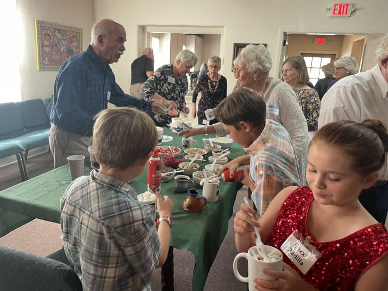 People gather around a table with a green tablecloth, enjoying an assortment of food items in a community setting.
