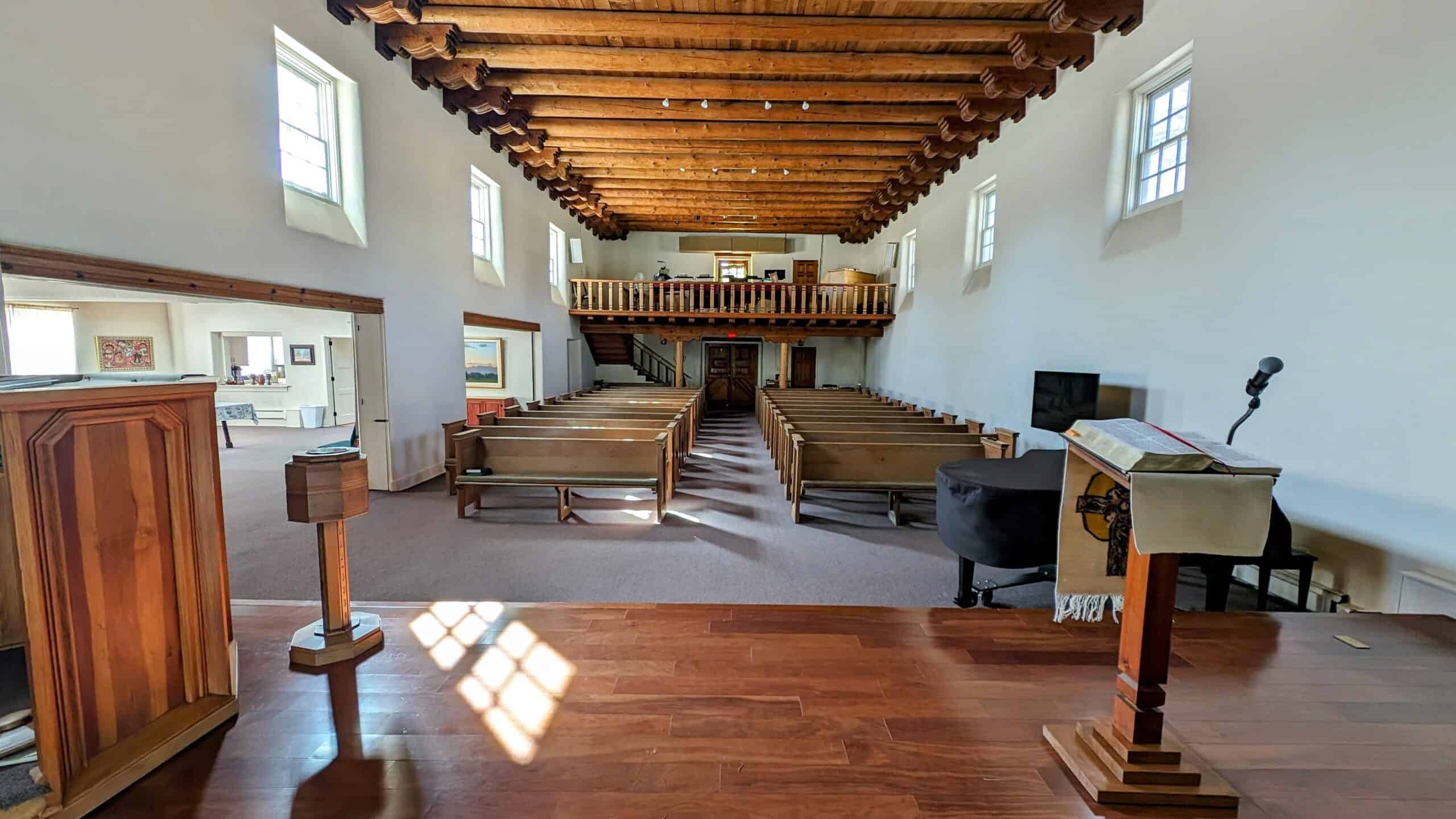 A wooden pulpit faces empty pews inside Albuquerque Friends Meetinghouse, with natural lighting from large windows. Historic beams line the ceiling.