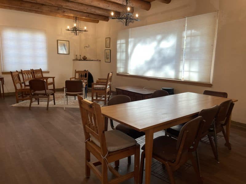 A cozy room features wooden tables and chairs, decorative light fixtures, and window blinds filtering soft light, creating a warm, inviting atmosphere.