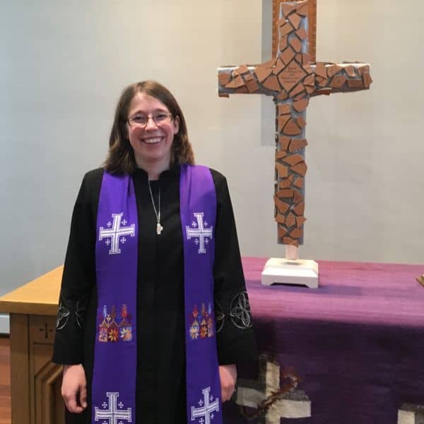 A person in religious attire stands smiling beside a decorative cross inside a church setting, with a purple draped altar.