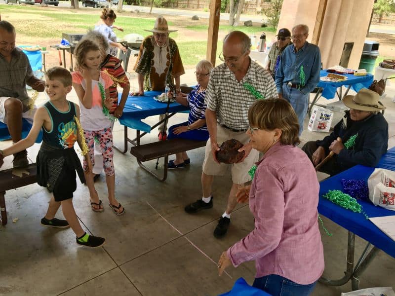 A group of people, including children, gather at a picnic shelter playing a game, surrounded by blue-covered tables and bags.