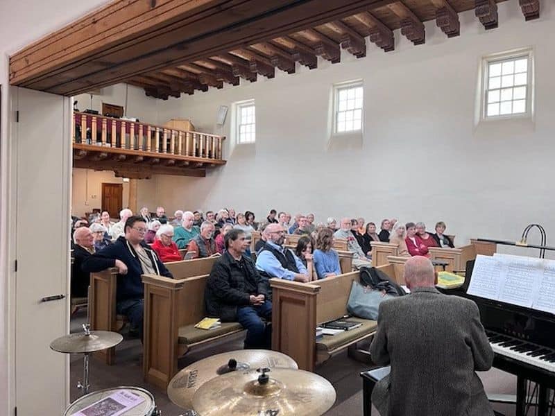 A group of people seated in a church, listening to a pianist. Wooden beams overhead, simple interior design, and an organ visible.