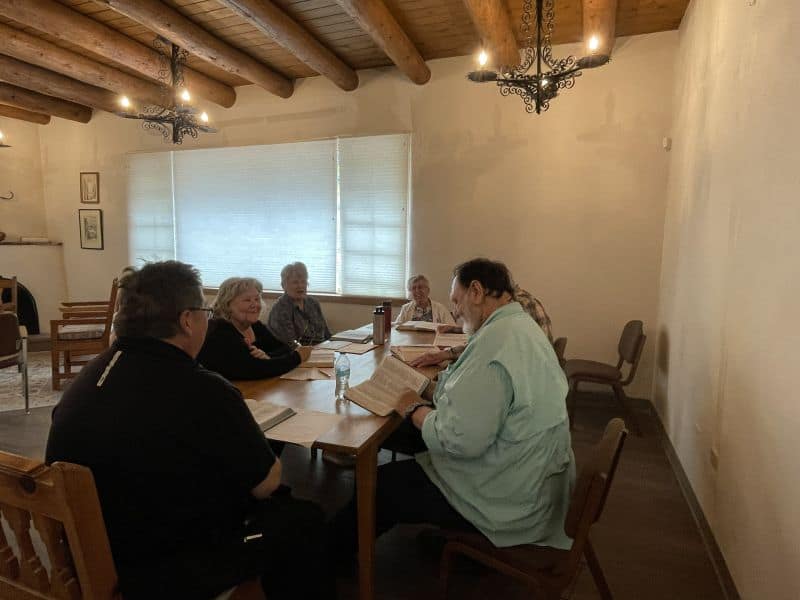 A group of people are gathered around a wooden table in a room with wooden beams and chandeliers, engaging in discussion.