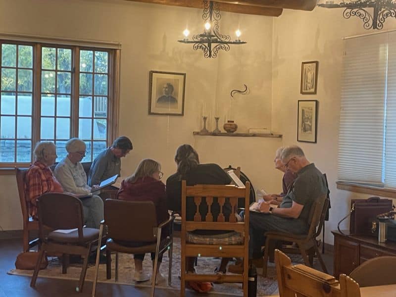 Seven people sit in a cozy, warmly lit room, engaged in discussion or study, surrounded by framed pictures and rustic decor.