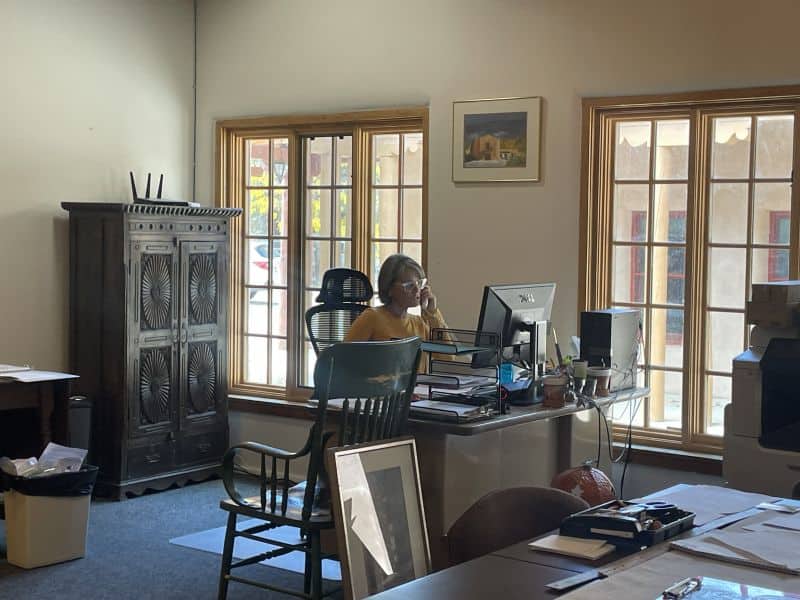 A person in an office on the phone, surrounded by desks, papers, and art. Large windows provide natural light to the room.