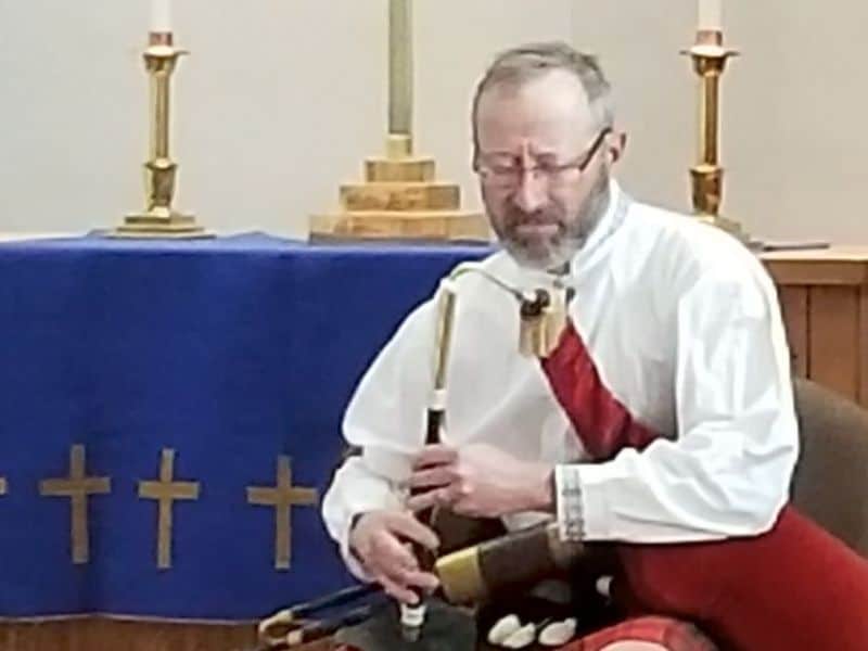 A person is playing bagpipes indoors, dressed in traditional attire. In the background, an altar with candlesticks and a cross-covered cloth is visible.