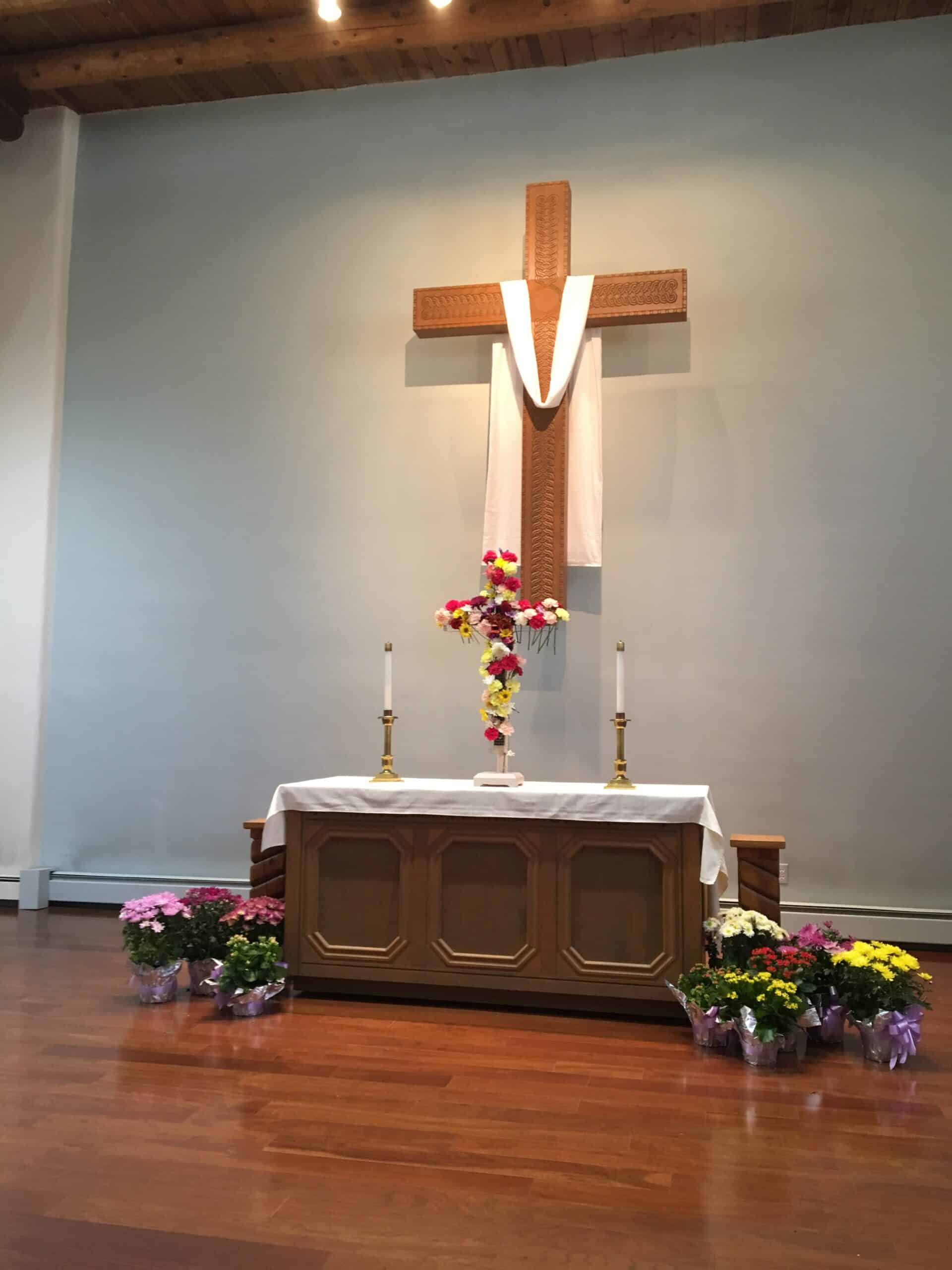 An altar with a wooden cross, draped fabric, colorful flowers, and candles in a church setting on a wooden floor.