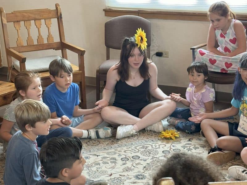 A person and children seated in a circle on a patterned rug, holding hands, with wooden chairs in the background.