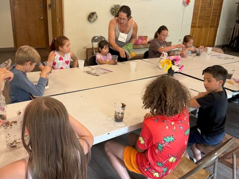 A person guides children seated around a table, engaging in a group activity with cups and materials on a white tabletop indoors.