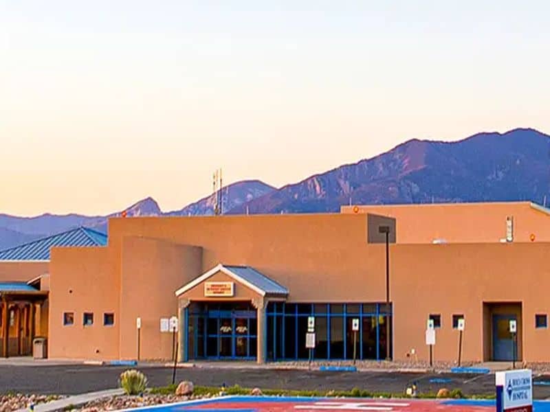 A beige stucco building with blue-framed windows sits in front of a mountain range under a clear sky during sunset.