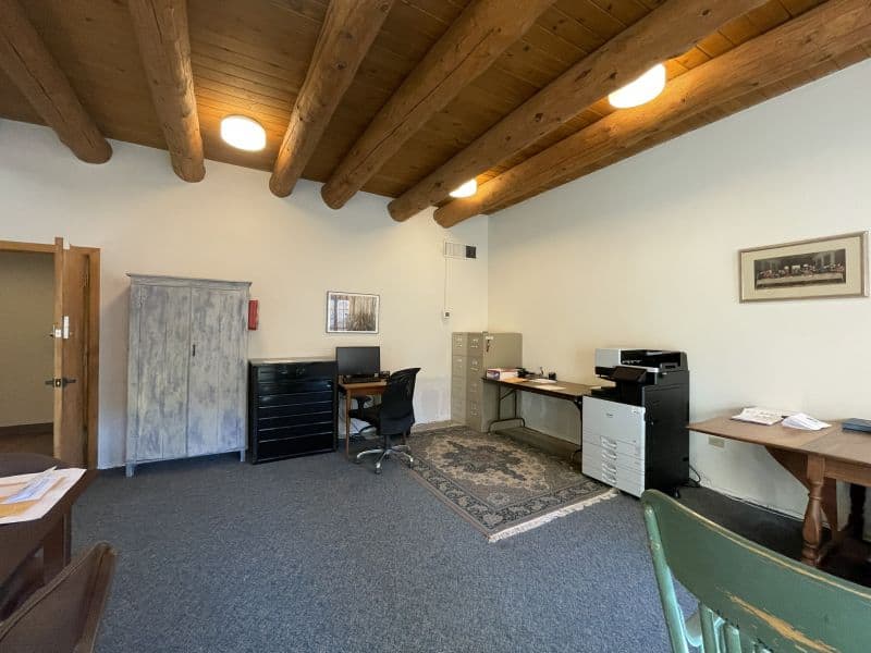A cozy office with wooden beams, desk, chairs, filing cabinets, printer, and framed pictures on walls, creating a warm and functional workspace.