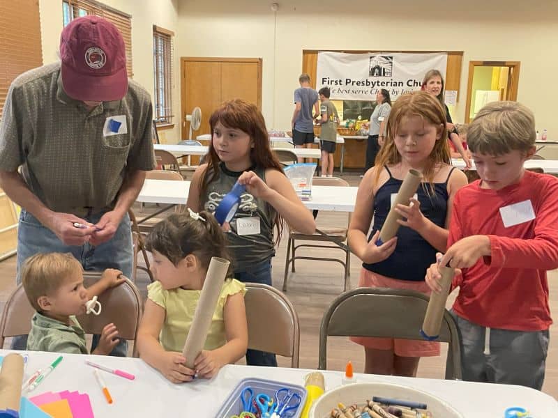 People and children engaged in crafting activity at First Presbyterian Church. Materials like cardboard tubes, markers, and scissors are on the table.