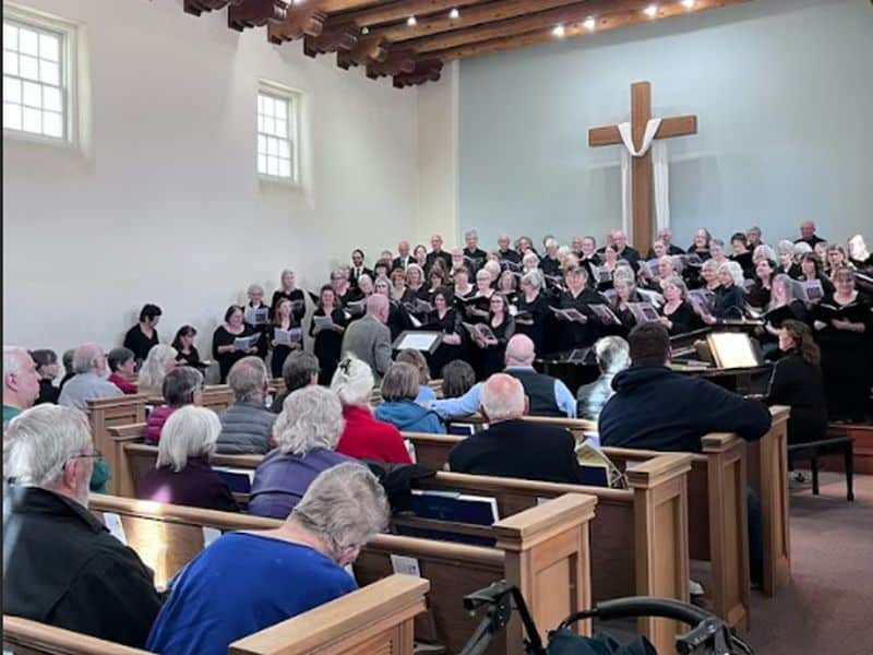 A choir performs in a church. People sit in pews listening. A large wooden cross is displayed at the front.