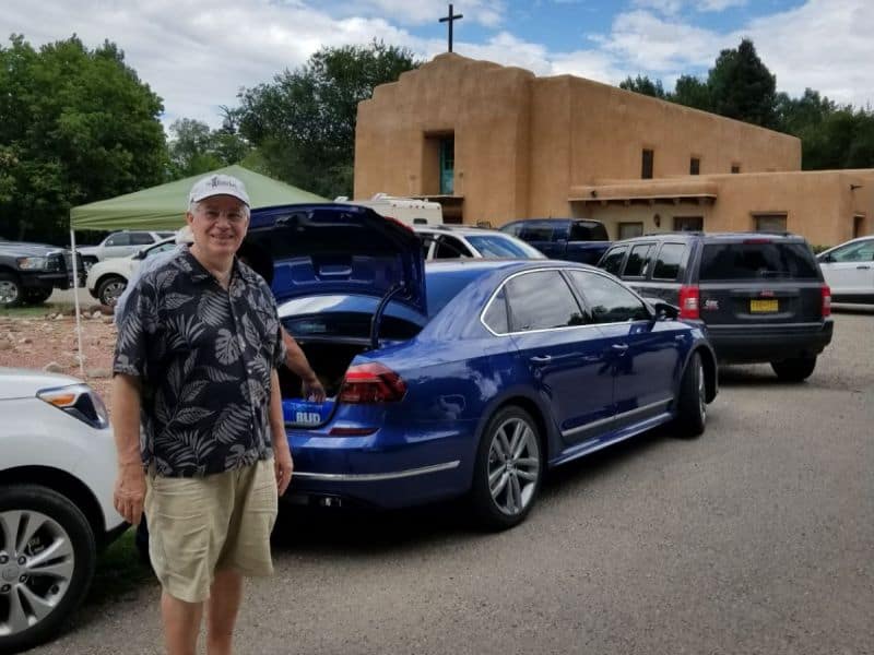A person stands near a blue car with an open trunk. An adobe church with a cross is visible in the background.
