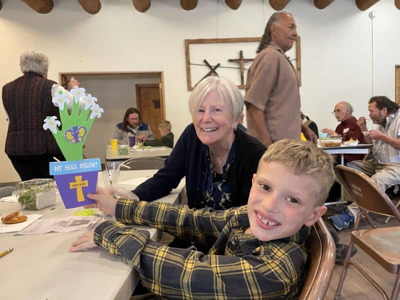 A person and child smile at a table displaying an Easter craft. People converse casually in a room with a cross on the wall.